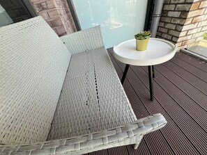 A wicker sofa with a white cushion, a small round table with a potted plant, and a view of a brick wall and window. - The Lyall - 1 Bedroom (London)