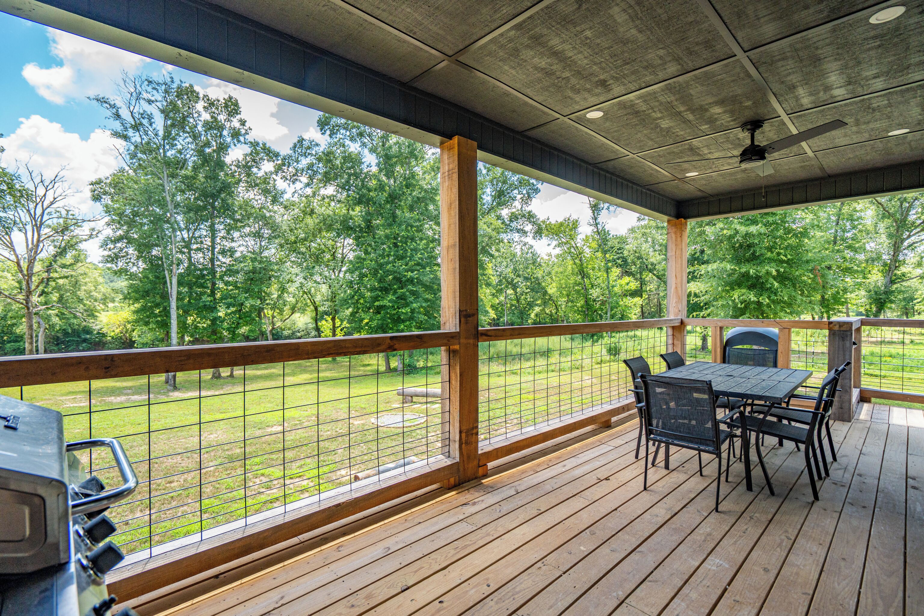 A covered outdoor deck with a table and chairs, a grill, and a view of a grassy area with trees.