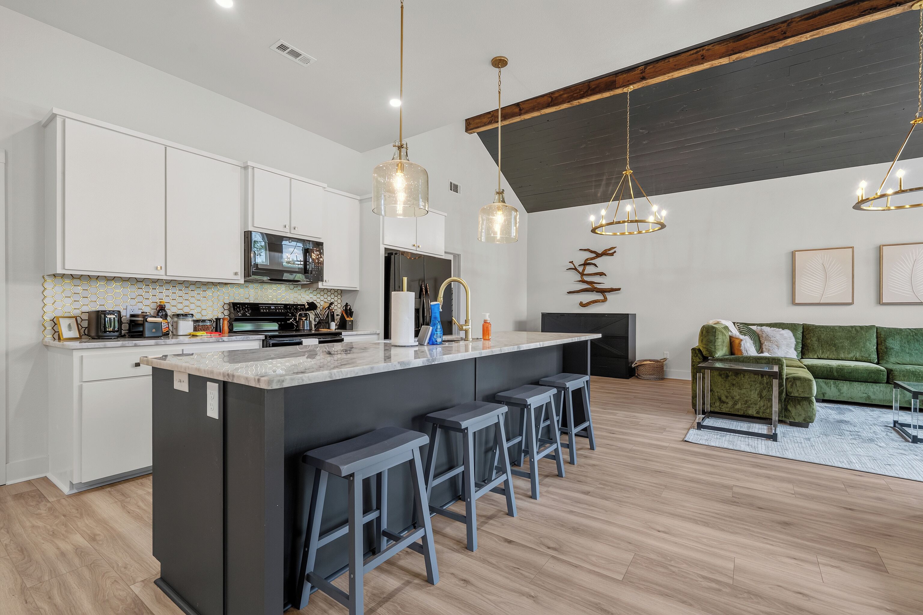 A modern kitchen with a central island, bar stools, pendant lighting, and a green sofa in a living area.