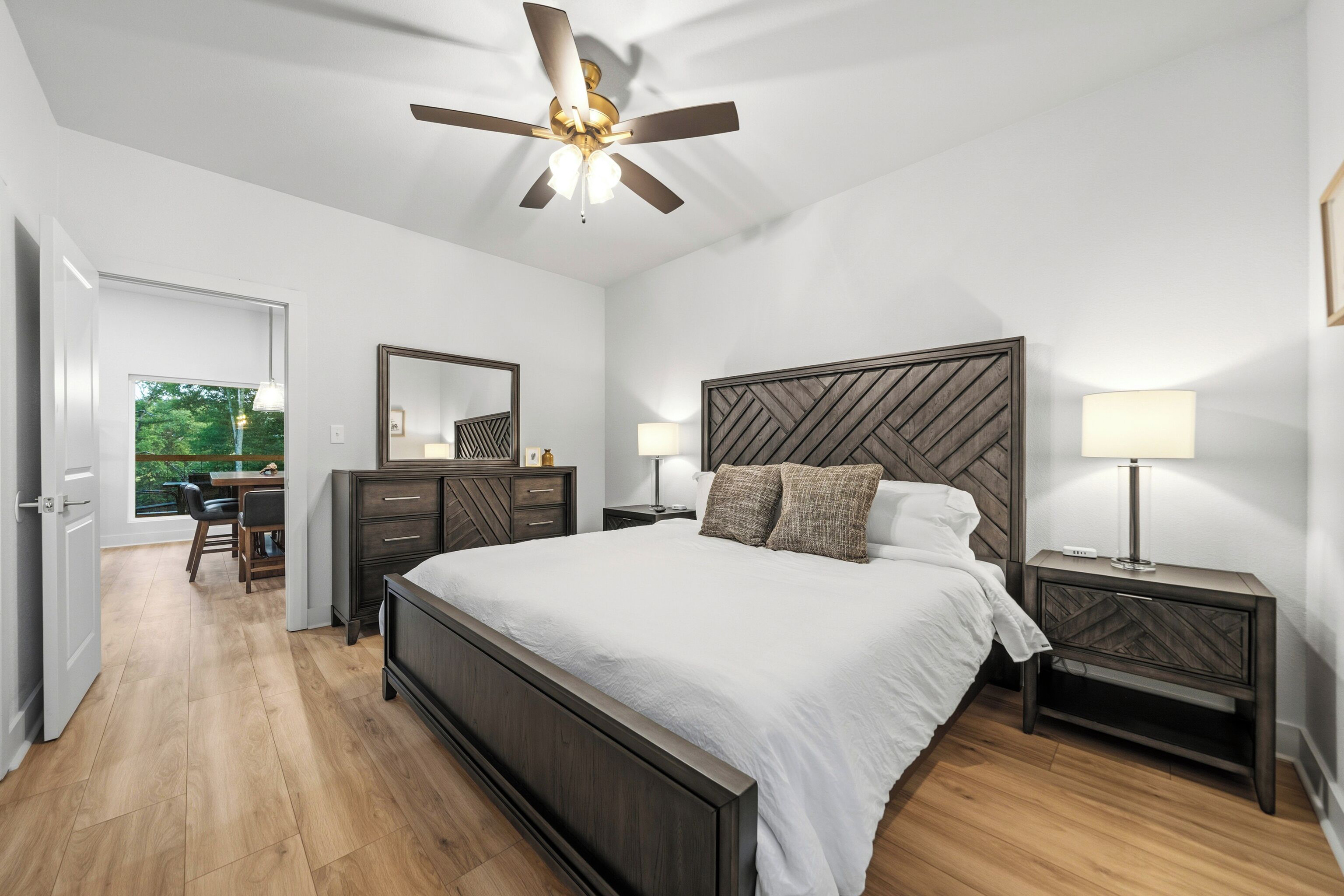Une chambre à coucher comprenant un lit, des tables de chevet, un ventilateur de plafond, une commode, un miroir et une vue sur l’extérieur.