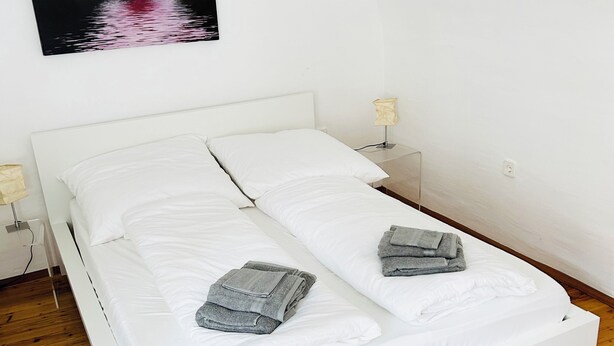 Habitación con cama individual, ropa de cama blanca, dos almohadas, una toalla doblada, mesita de noche con lámpara y un cuadro de flores rosadas colgado en la pared.