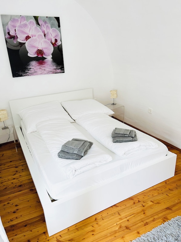Une chambre avec un lit simple, un couvre-lit blanc, deux oreillers, une serviette pliée, une table de chevet avec une lampe et un tableau représentant des fleurs roses accroché au mur.