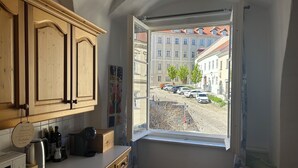 Una habitación con cocina, una ventana que muestra coches aparcados y edificios, y una cama con un cojín rojo.