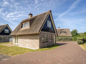 Een woonhuis met een gevel van baksteen, een rieten dak en een geplaveid pad dat leidt naar een poort.