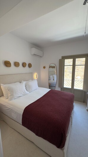 A bedroom with a bed, a wall-mounted air conditioner, a mirror, and a window with a view. - Borgo Stampalia – Villa Terracotta (Astypalea)