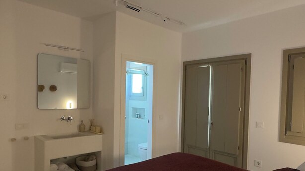 A bedroom with a bed, a bedside table, a mirror, a wardrobe, and a bathroom visible through an open door. - Borgo Stampalia – Villa Terracotta (Astypalea)