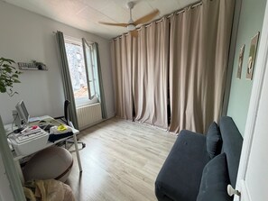 Ein Zimmer mit Schreibtisch, Stuhl und einer Couch. Ein Fenster mit Vorhängen, ein Deckenventilator und eine Heizung sind ebenfalls vorhanden.
