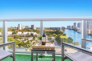 A balcony with a table, two chairs, a bottle of wine, and a glass. A cityscape and river are visible in the background. - Beachwalk- Studio with Pool & Bay View (Hallandale Beach)