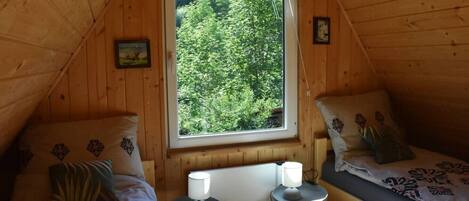 A cozy attic bedroom with two beds, a window with a forest view, and a small table with a lamp.