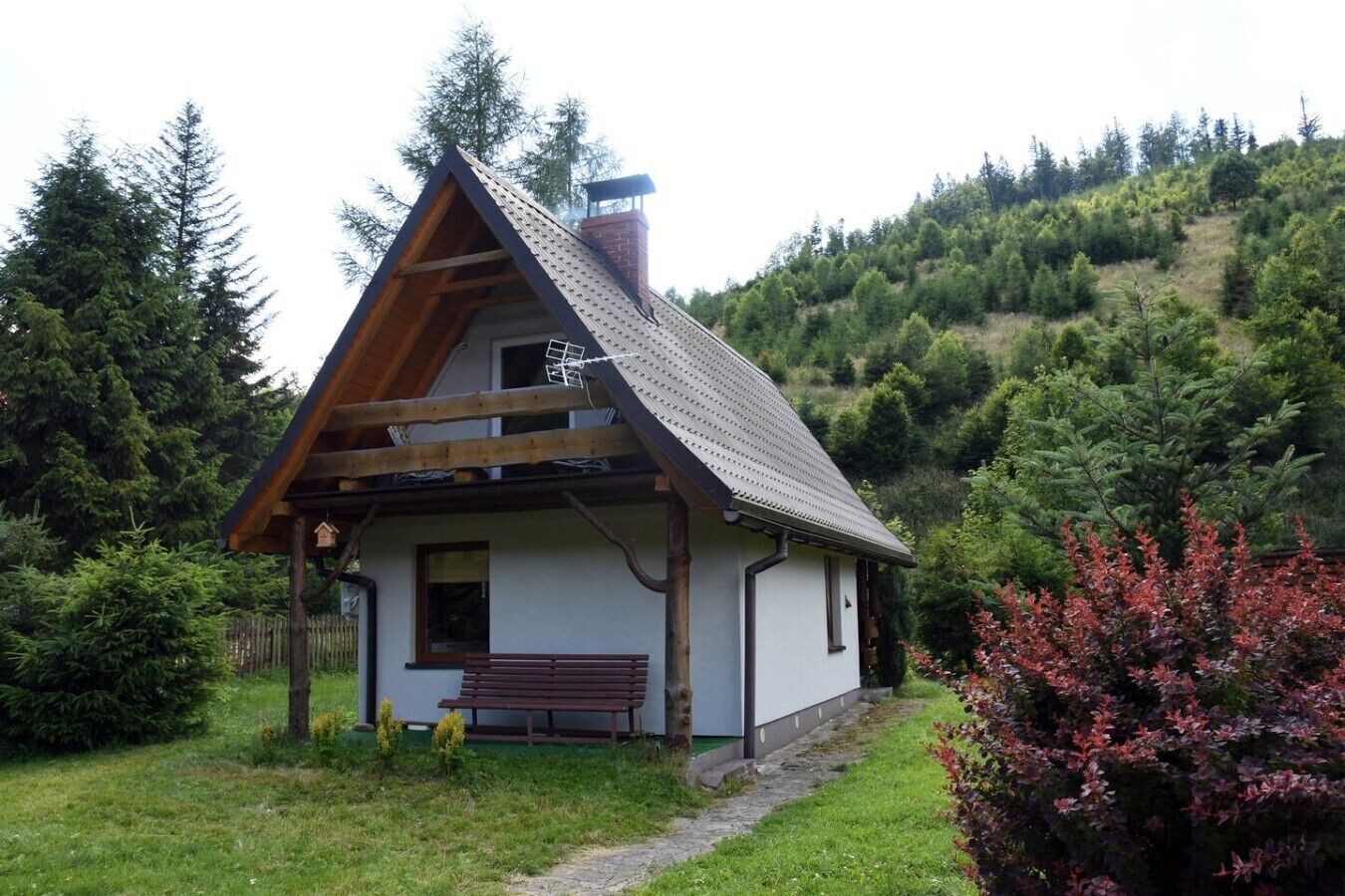 A small house with a sloped roof, wooden beams, and a chimney. It is surrounded by greenery and a bench is visible in front.