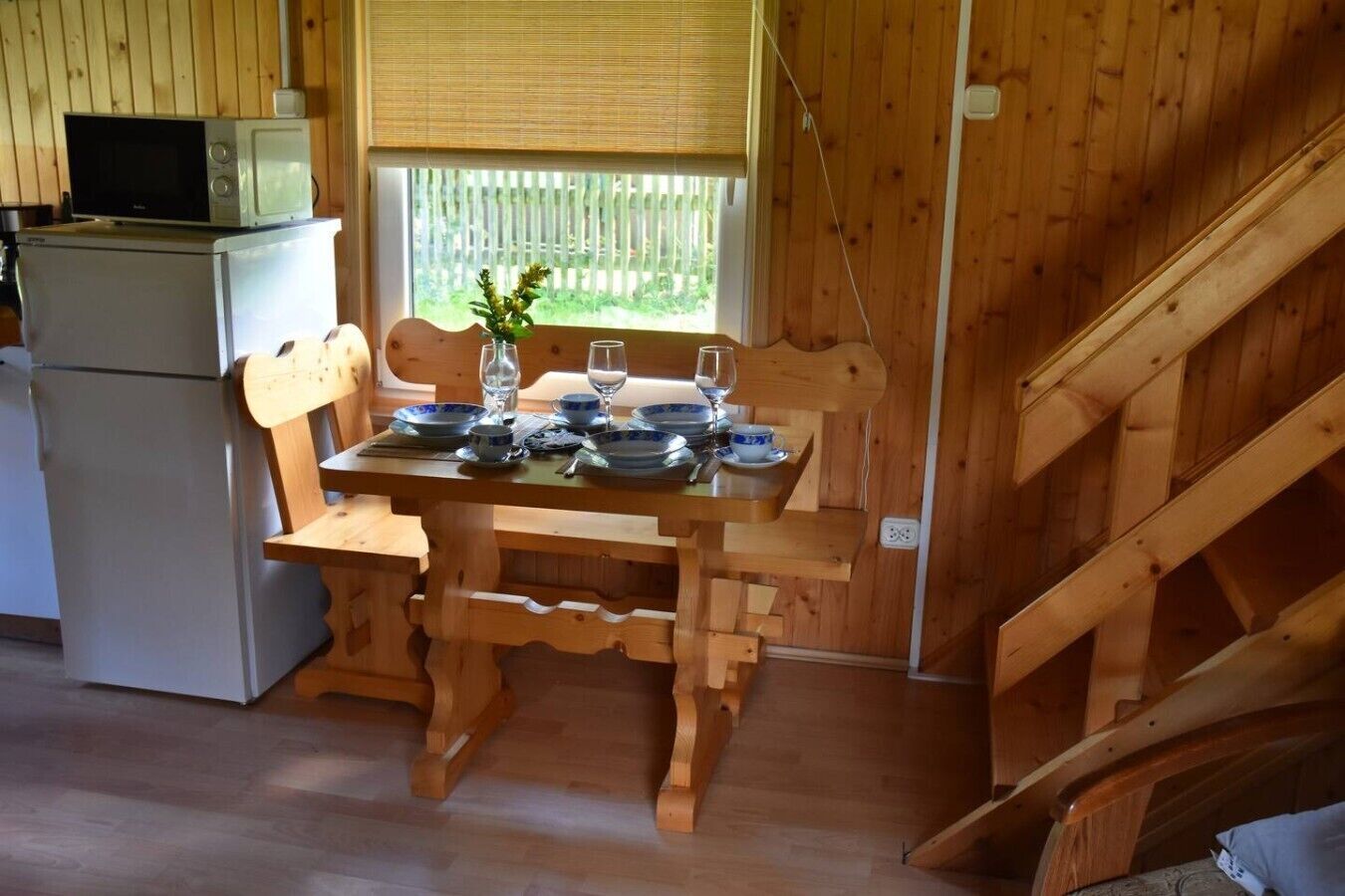 Een houten eettafel gedekt met borden, glazen en een vaas bloemen. Een magnetron staat op een klein tafeltje links.