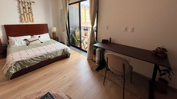A hotel room with a bed, a desk, a chair, a window with curtains, and a balcony with plants. - Mexican Designer Studio - Pool, Gym, and Rooftop (Ciudad de México)