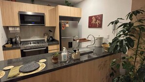 A modern kitchen with wooden cabinets, a stainless steel microwave, a gas stove, and a refrigerator. There is a bar area with stools and a potted plant. - Mexican Designer Studio - Pool, Gym, and Rooftop (Ciudad de México)
