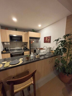 A modern kitchen with wooden cabinets, a stainless steel microwave, a gas stove, and a refrigerator. There is a bar area with stools and a potted plant. - Mexican Designer Studio - Pool, Gym, and Rooftop (Ciudad de México)