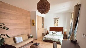 A modern bedroom with a wooden headboard bed, a sitting area with a sofa and chairs, a dining table, and a hanging light fixture. - Mexican Designer Studio - Pool, Gym, and Rooftop (Ciudad de México)