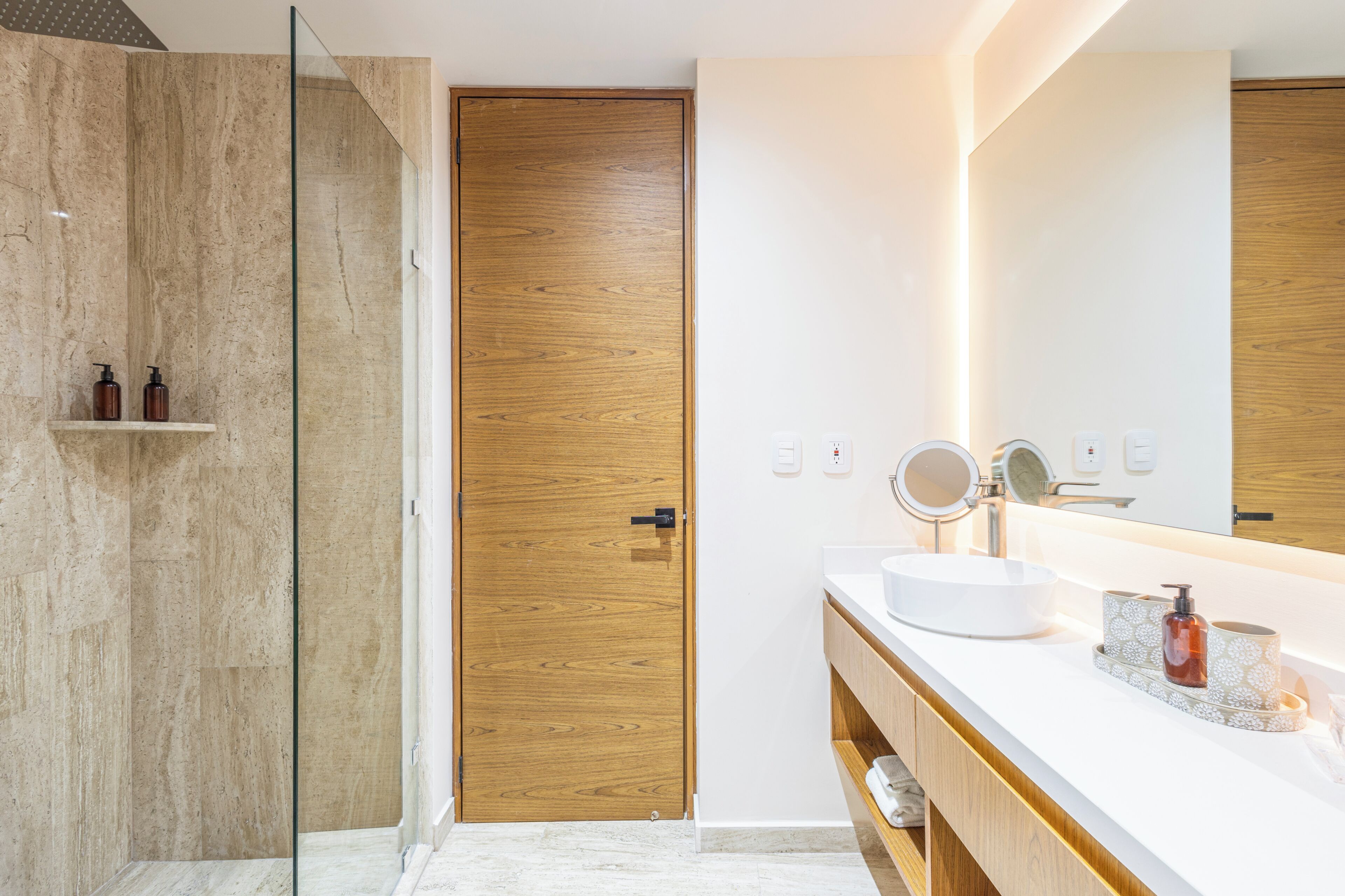 Ein modernes Badezimmer mit einer Duschkabine aus Glas, einer Holztür, einem weißen Waschbecken mit rundem Spiegel und einer Holzkommode.