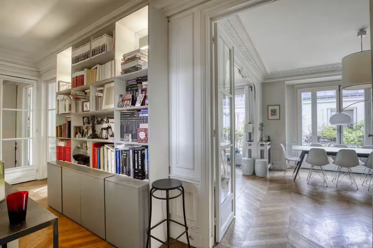 Een ruime kamer met een grote boekenkast, een zithoek met stoelen, en een barkruk.