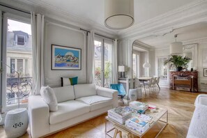 Ein geräumiges Wohnzimmer mit einem weißen Sofa, einem Couchtisch mit Büchern, einem Gemälde an der Wand und einem Balkon mit Geländer.