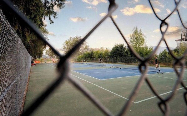 Un court de tennis entouré d’une clôture en treillis métallique, avec des arbres et un ciel dégagé.