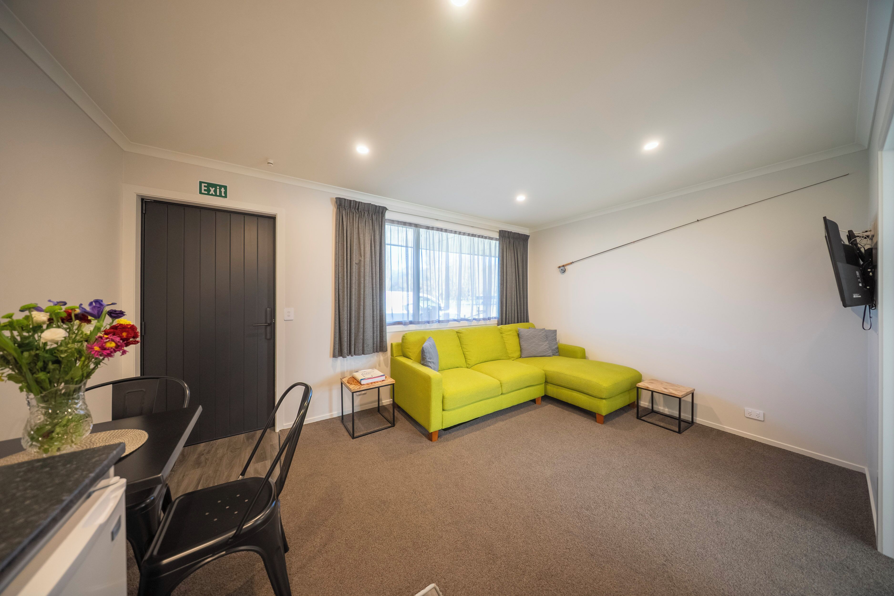 A modern living room with a green sofa, a dining table with chairs, a small side table, and a TV mounted on the wall.