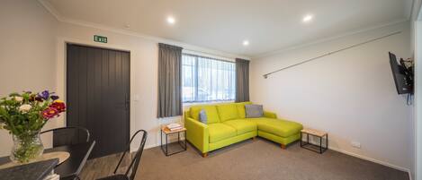 A modern living room with a green sofa, a dining table with chairs, a small side table, and a TV mounted on the wall.