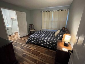 Un dormitorio con cama, una silla, un escritorio con lámpara y una puerta que da a un baño.