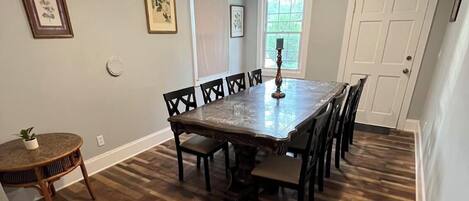 A dining room with a large rectangular table surrounded by chairs, a small round table with a plant, and a window with blinds.