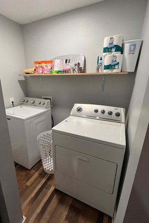 A laundry room with two washing machines, a dryer, and a shelf with laundry supplies.