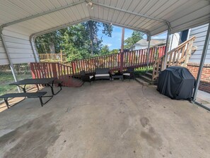 Un patio techado al aire libre con piso de concreto, una terraza de madera con barandal rojo y una parrilla cubierta.