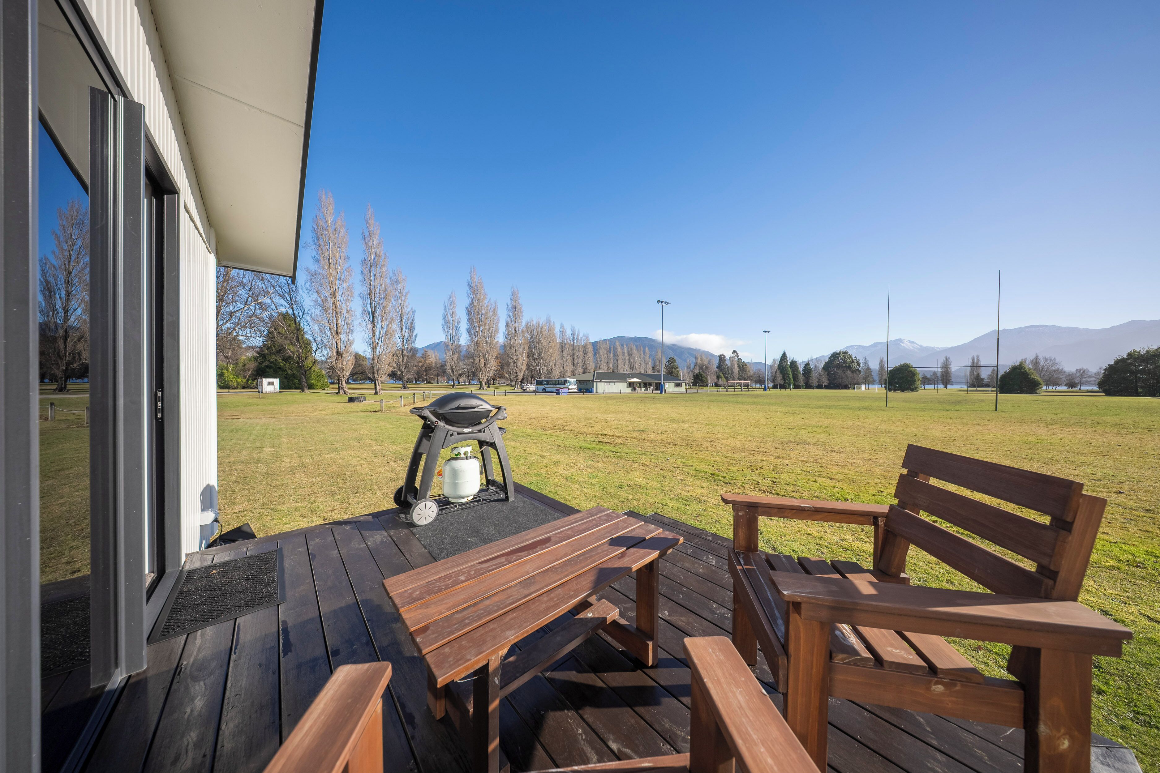 A patio with wooden furniture, a grill, and a view of a grassy field and mountains.