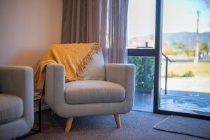 Ein Zimmer mit einem grauen Sessel, der mit einer gemusterten Decke bedeckt ist, einem Fenster mit Vorhängen und Blick auf ein Gebäude sowie Grünflächen.