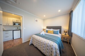 A bedroom with a bed, bedside table, lamp, and a view of a kitchenette with a microwave. - Dusky Mustering Unit - Te Anau Holiday Home (Te Anau)