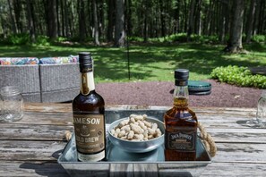 Zwei Whiskeyflaschen, eine Schüssel Erdnüsse und ein Glas auf einem Holztisch im Freien.