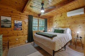 Un dormitorio tipo cabaña de madera con una cama, un ventilador de techo, una ventana y aire acondicionado de pared.