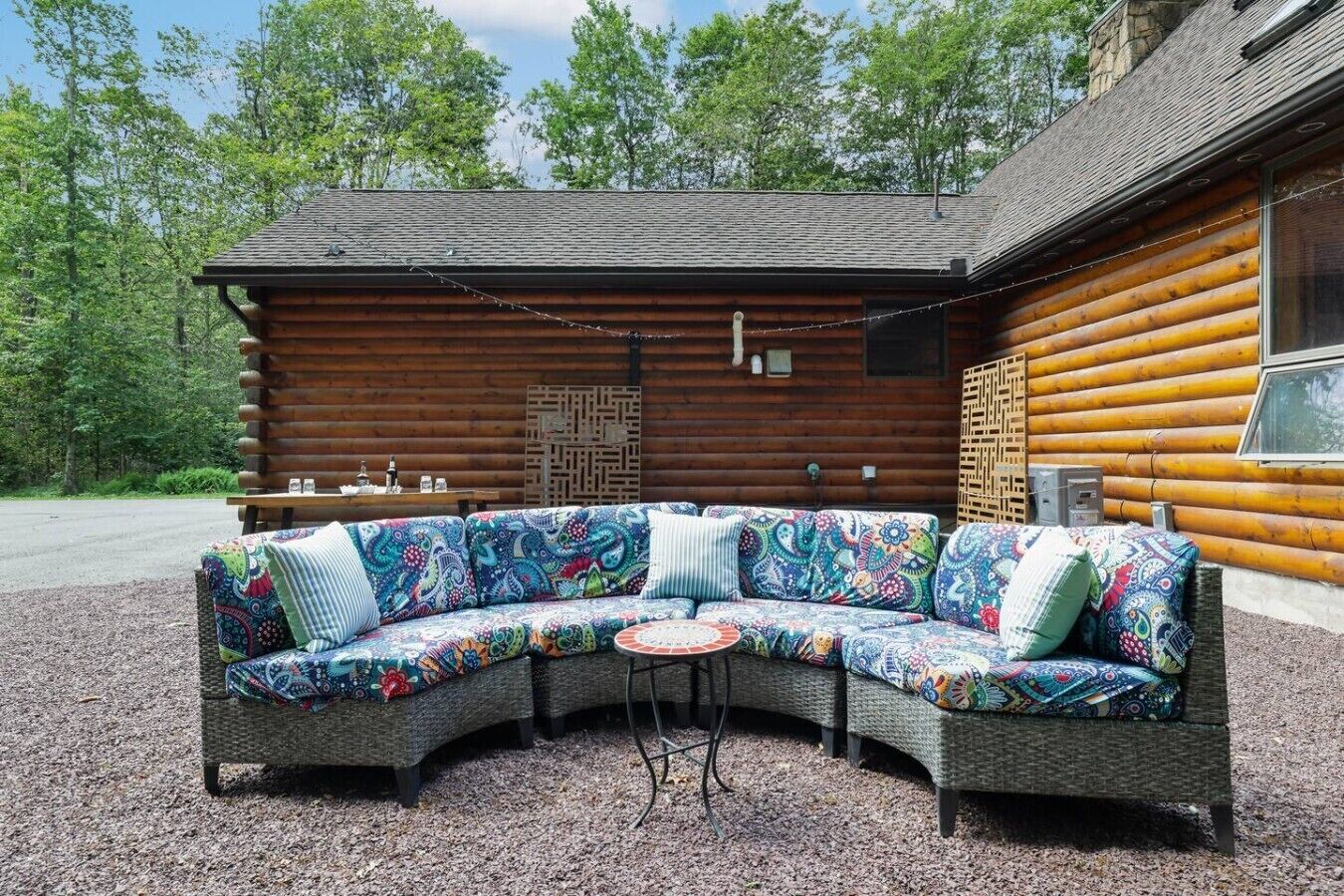 Une cabane en rondins dotée d’un espace extérieur aménagé avec des sièges incurvés, comprenant un canapé aux motifs colorés et une petite table ronde.