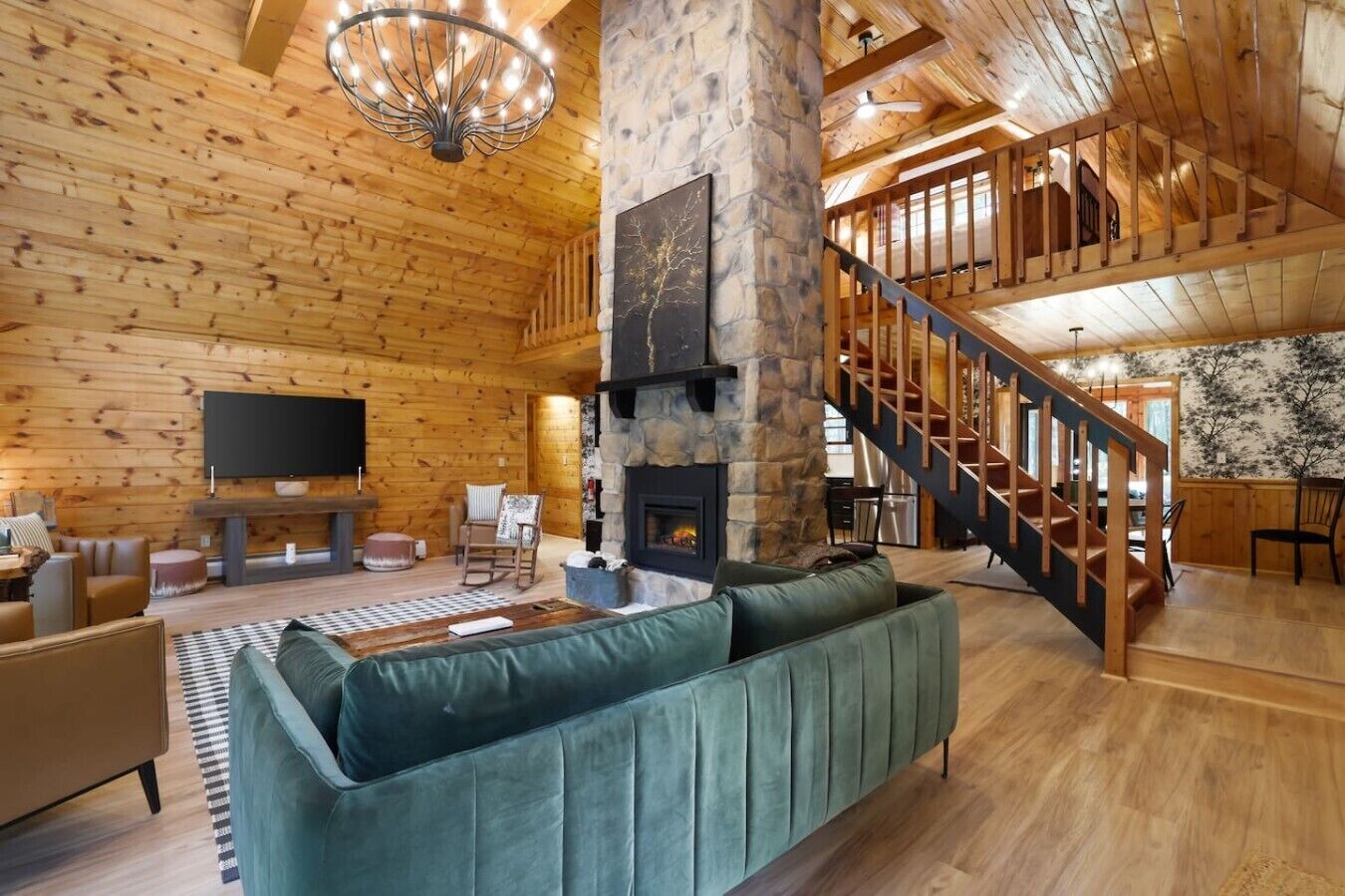 Un vaste espace de vie agrémenté d’une cheminée en pierre, d’un grand canapé vert, de parquets en bois et d’un escalier menant à un niveau supérieur.