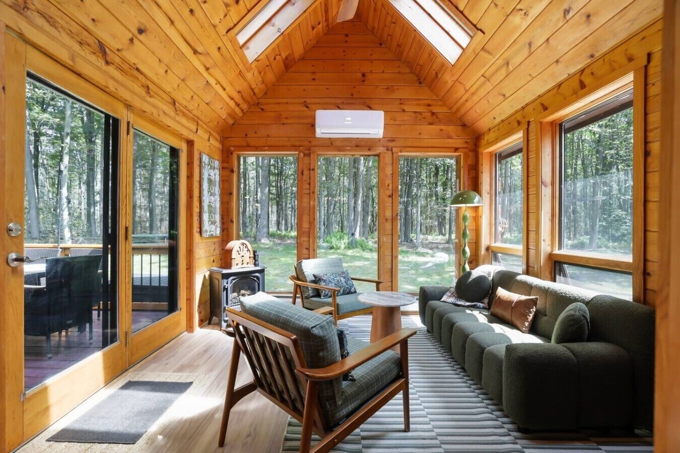 A log cabin interior with a sofa, chairs, a fireplace, and large windows offering forest views.