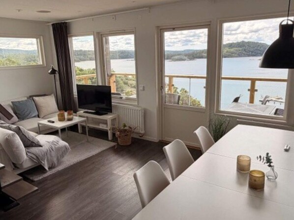 Una sala moderna con sofá, televisión y comedor. Amplias ventanas ofrecen vistas a un lago y colinas.
