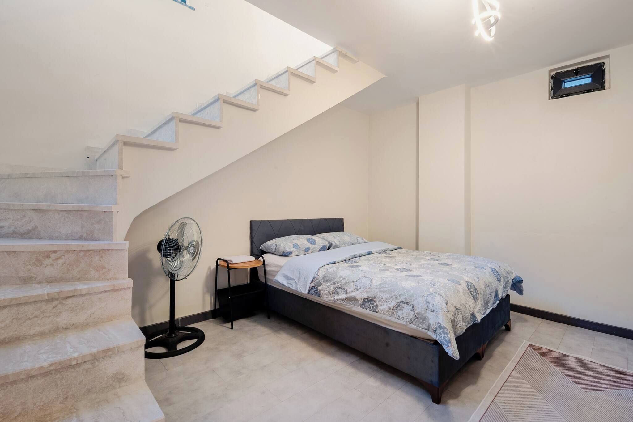 A bedroom with a bed, a standing fan, a small table, a staircase, and a wall-mounted air vent.