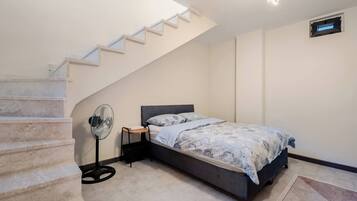 A bedroom with a bed, a standing fan, a small table, a staircase, and a wall-mounted air vent.