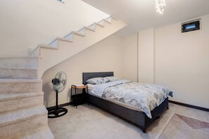 A bedroom with a bed, a standing fan, a small table, a staircase, and a wall-mounted air vent.