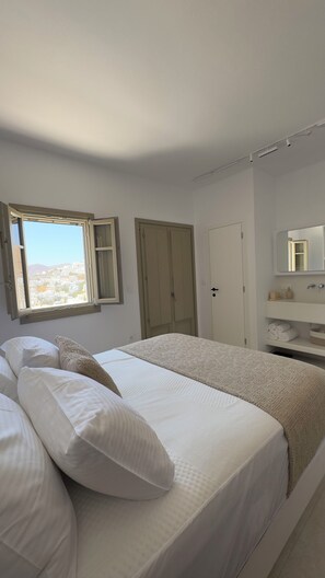 A bedroom with a bed, a wardrobe, a bathroom with a mirror, and a window with a view. - Borgo Stampalia – Villa Sabbia (Astypalea)