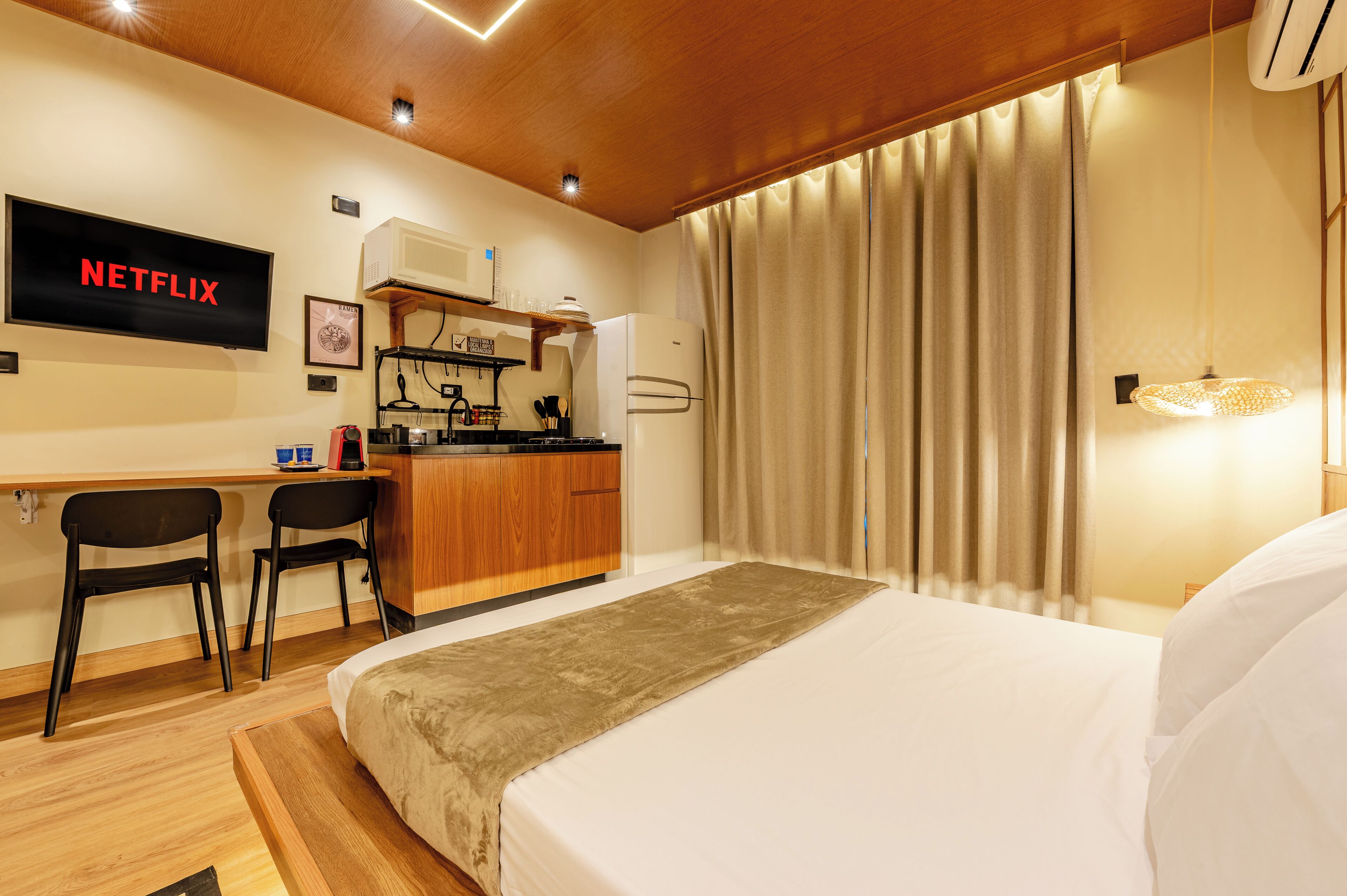 A hotel room with a bed, a desk, a chair, a kitchenette, and a TV screen displaying 'NETFLIX'.