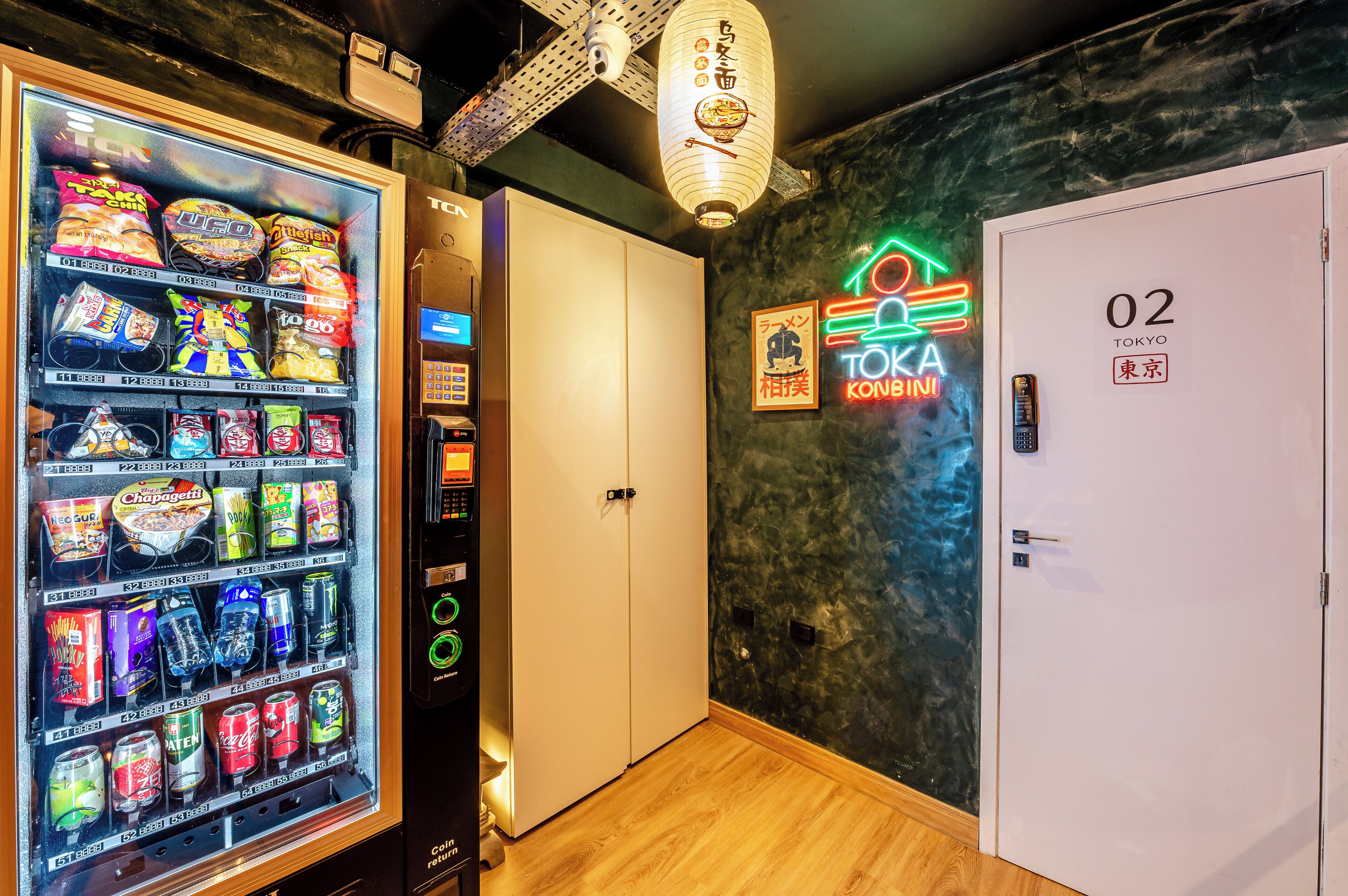 Máquina expendedora con aperitivos y bebidas, un letrero de neón que dice 'TOKA KOBIN' y una puerta con la etiqueta '02 TOKYO'.