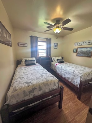 A room with two single beds, a ceiling fan, a window with curtains, and a wall sign indicating 'BOATS'. - 3-bedroom Waterview cabin with AC in charming Birchwood | Waterview Cabin with Private Boat Slip (Birchwood)