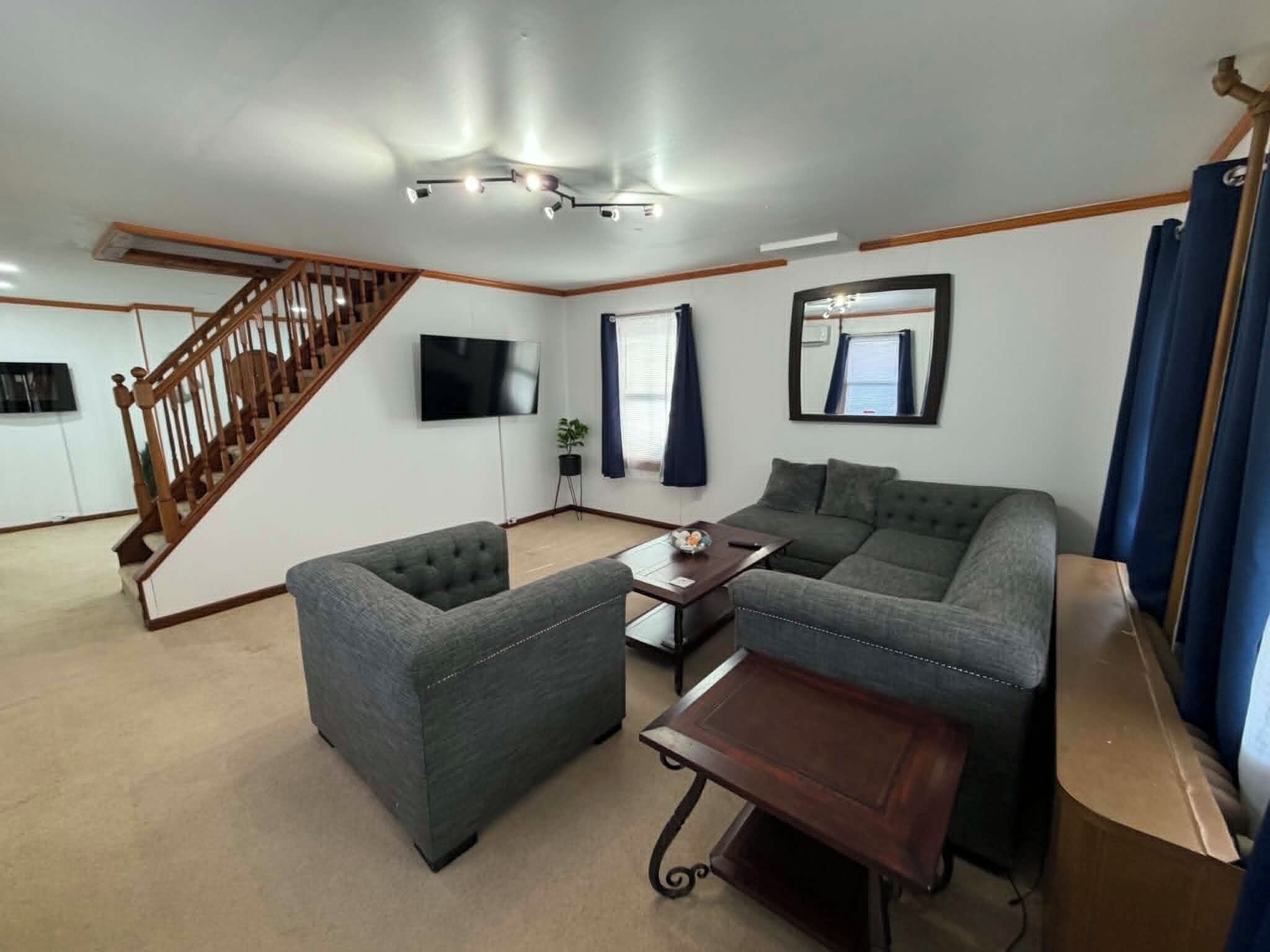 Ein Wohnzimmer mit Sofa, Hocker und einem kleinen Tisch. Eine Treppe führt in einen anderen Raum, ein Flachbildfernseher ist an der Wand montiert und ein Fenster mit Vorhängen vorhanden.
