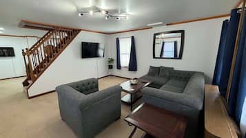 Ein Wohnzimmer mit Sofa, Hocker und einem kleinen Tisch. Eine Treppe führt in einen anderen Raum, ein Flachbildfernseher ist an der Wand montiert und ein Fenster mit Vorhängen vorhanden.