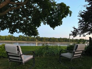 Two outdoor chairs facing a body of water with a tree overhead.