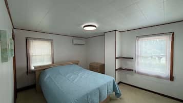 A bedroom with a bed, two windows with blinds, a bedside table, a small dresser, and an air conditioning unit.