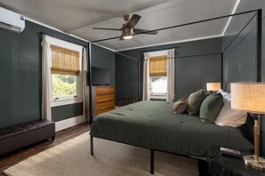 Ein Schlafzimmer mit einem Bett, Deckenventilator, einer Bank, einem Schminktisch und einem Fenster mit Jalousien.